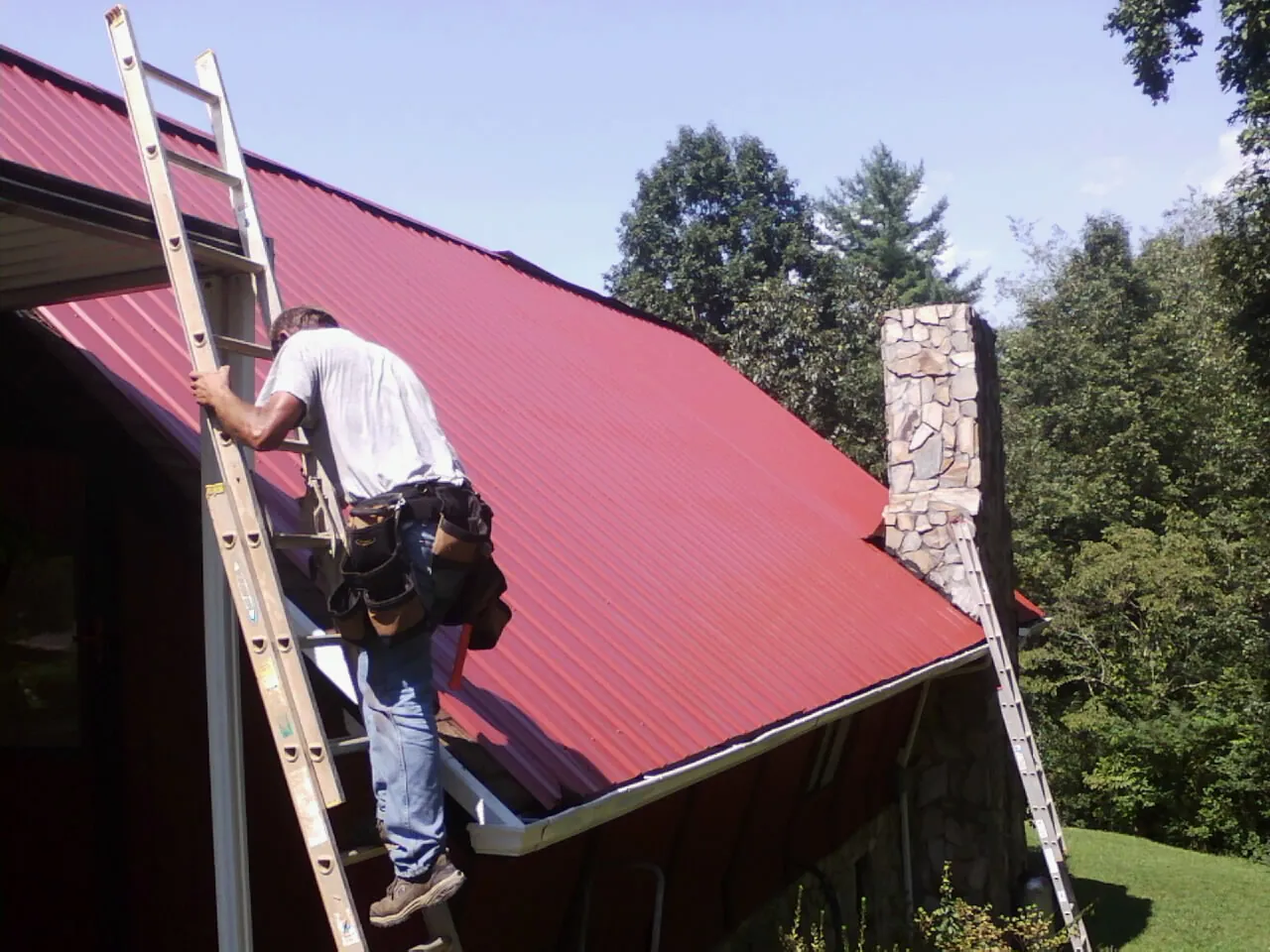 Roofer on a ladder beside a red metal roof and stone chimney — Soffit & Fascia Repair by Roofing Brothers in Groton